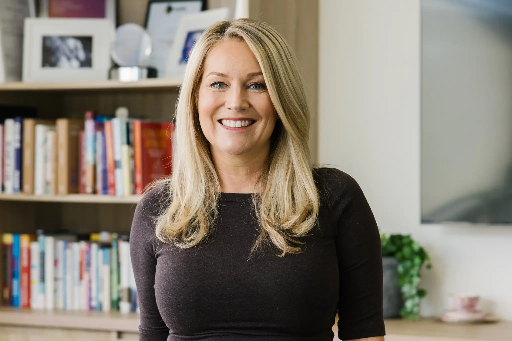 Tammy Barton, founder and director of MyBudget, smiling at the camera in a dark fitted top. She has shoulder-length blonde hair and is set against a brightly lit background featuring bookshelf shelves filled with books, awards, and framed photo frames.